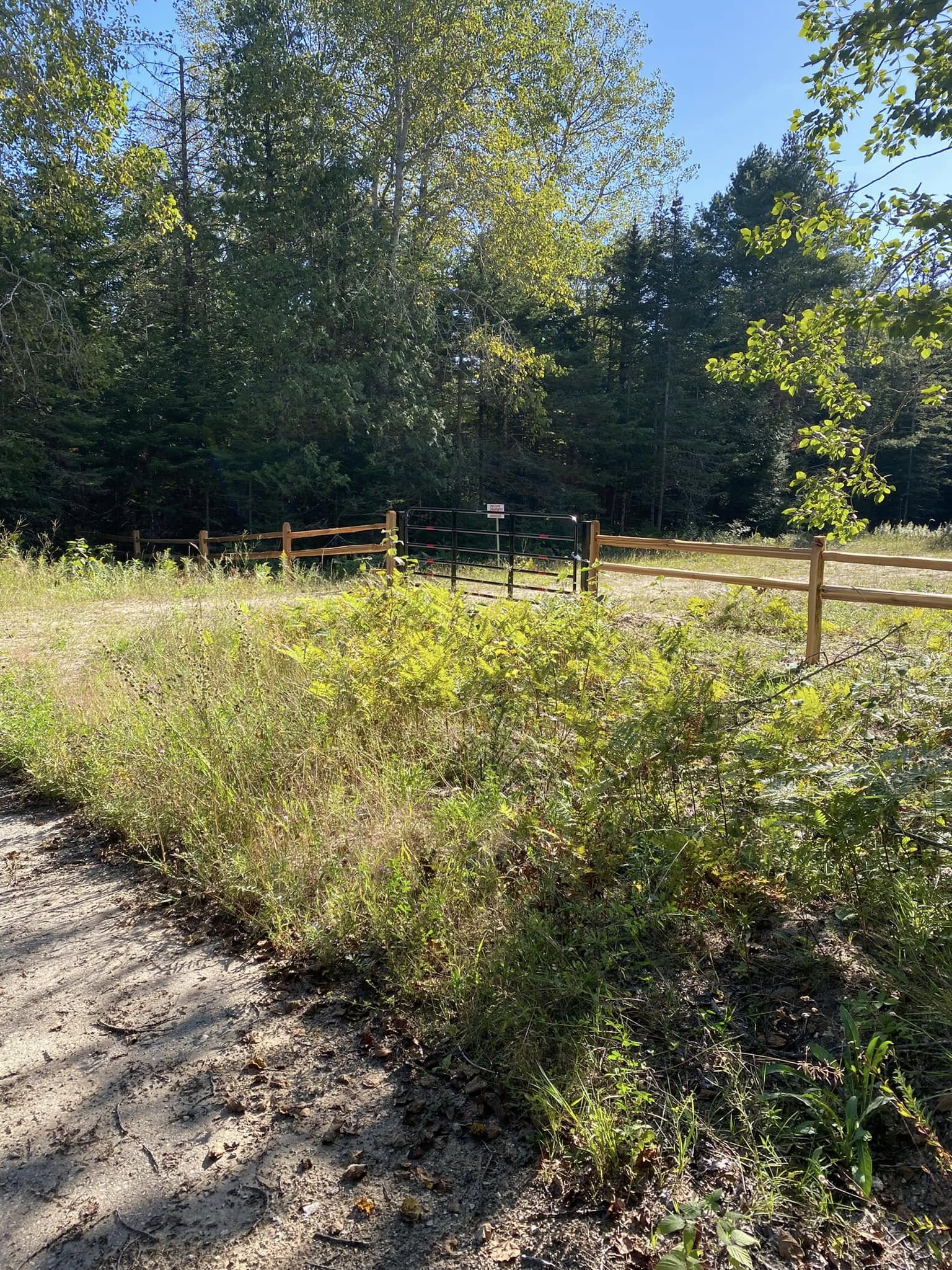 Cornerstone_9 Black metal farm gate between wooden fences and green trees.